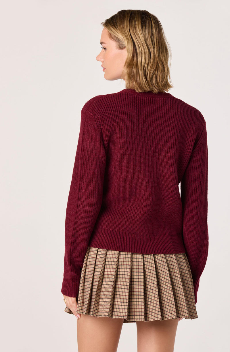 A person is wearing a maroon, long-sleeved, knitted sweater with a crew neckline. They are also wearing a pleated skirt with a beige and black checkered pattern.