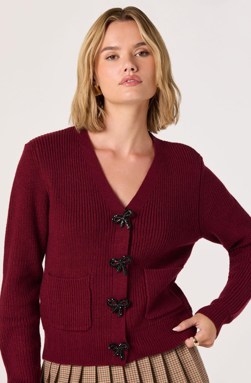 A woman is wearing a deep red, knitted cardigan with a V-neckline and four decorative black bows down the front. The cardigan has a textured ribbed pattern and two front patch pockets. She is also wearing a beige and brown checkered skirt.
