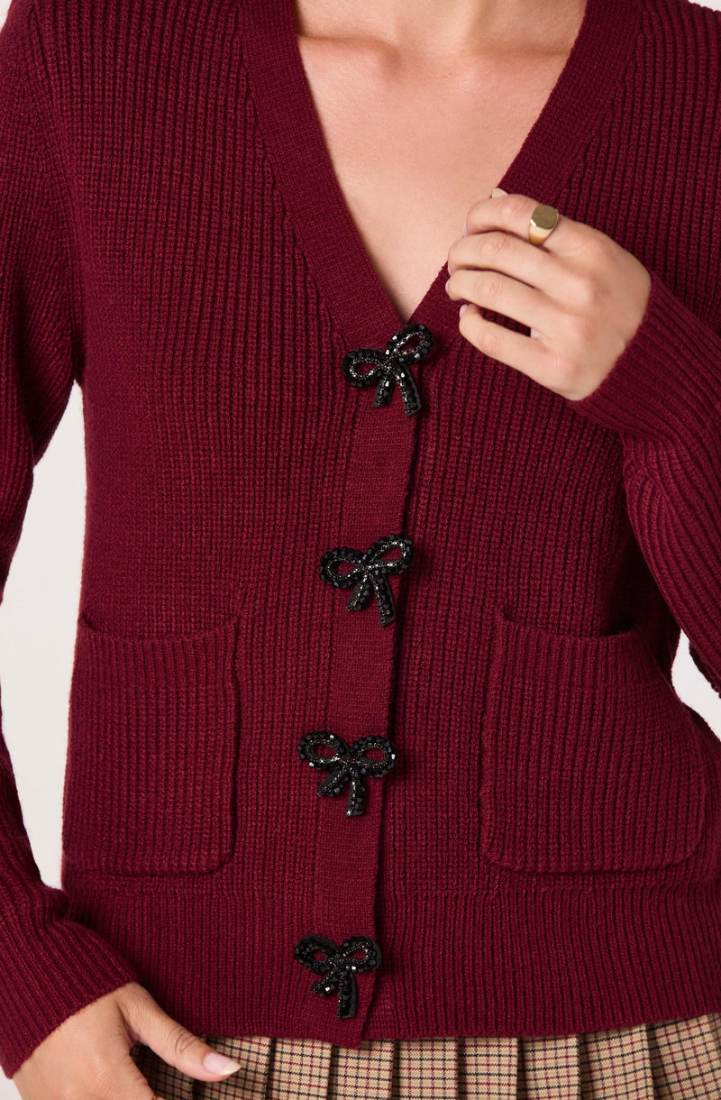 A person is wearing a deep red knit cardigan with a ribbed texture. The cardigan features a V-neckline and is fastened with four decorative black bows along the front opening. The person has their right hand near their collar, showcasing a simple gold ring. They are also wearing a plaid skirt with a beige and black pattern.