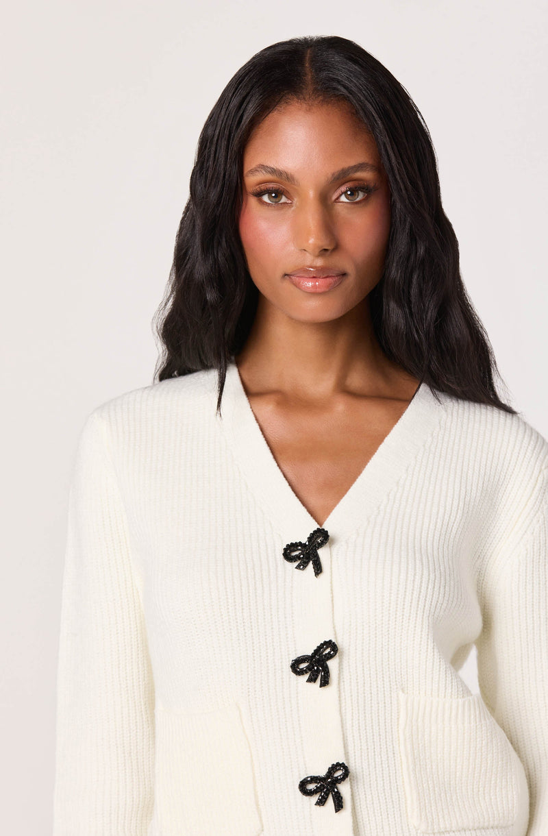 A woman wearing a cream-colored, ribbed knit cardigan with a v-neckline. The cardigan features three black bow-shaped buttons down the front.
