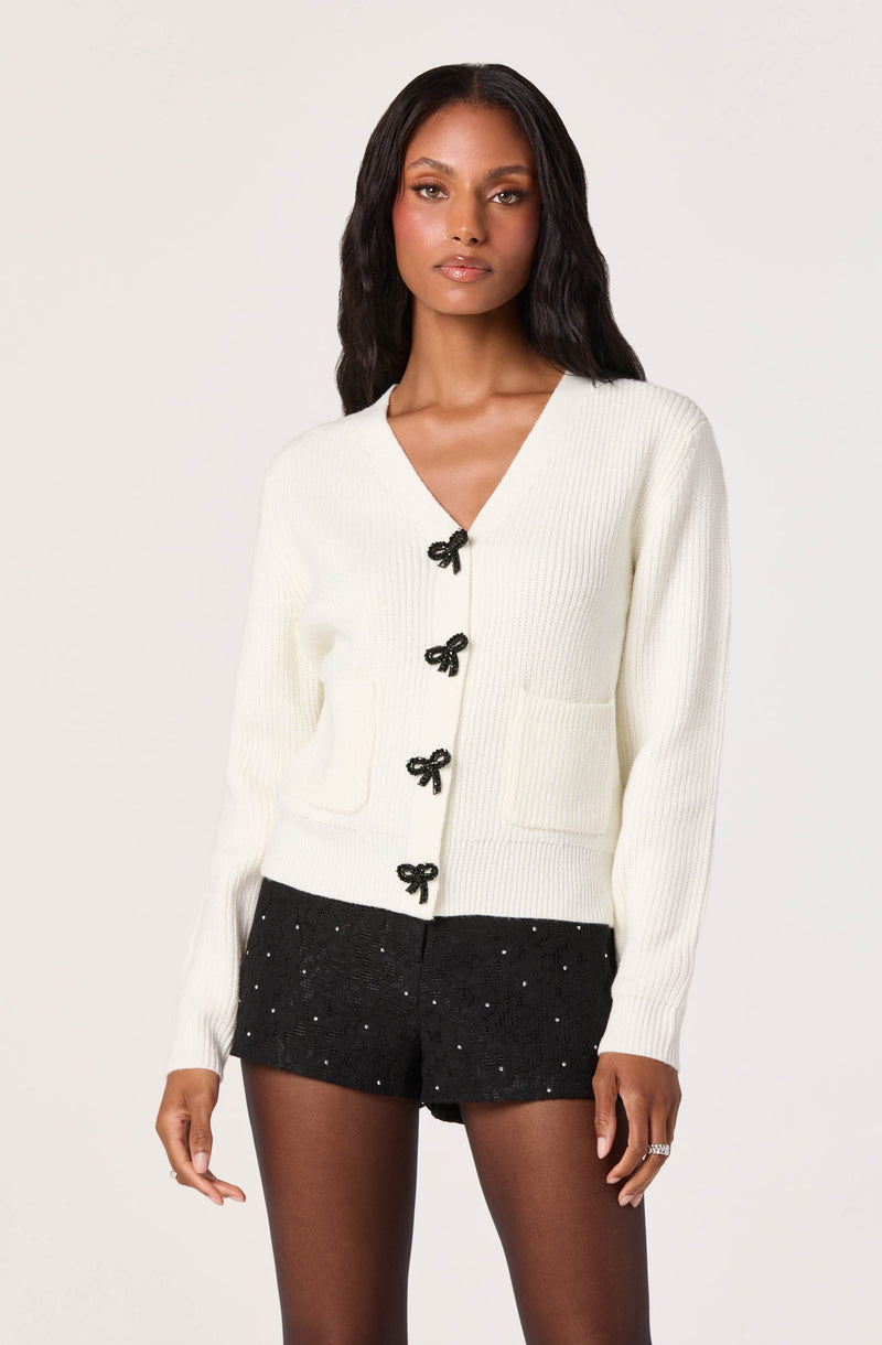 A woman is wearing a white, long-sleeved, V-neck knit sweater with black bow-shaped buttons down the front and two front pockets. She pairs it with black shorts that have small white polka dots or embellishments.