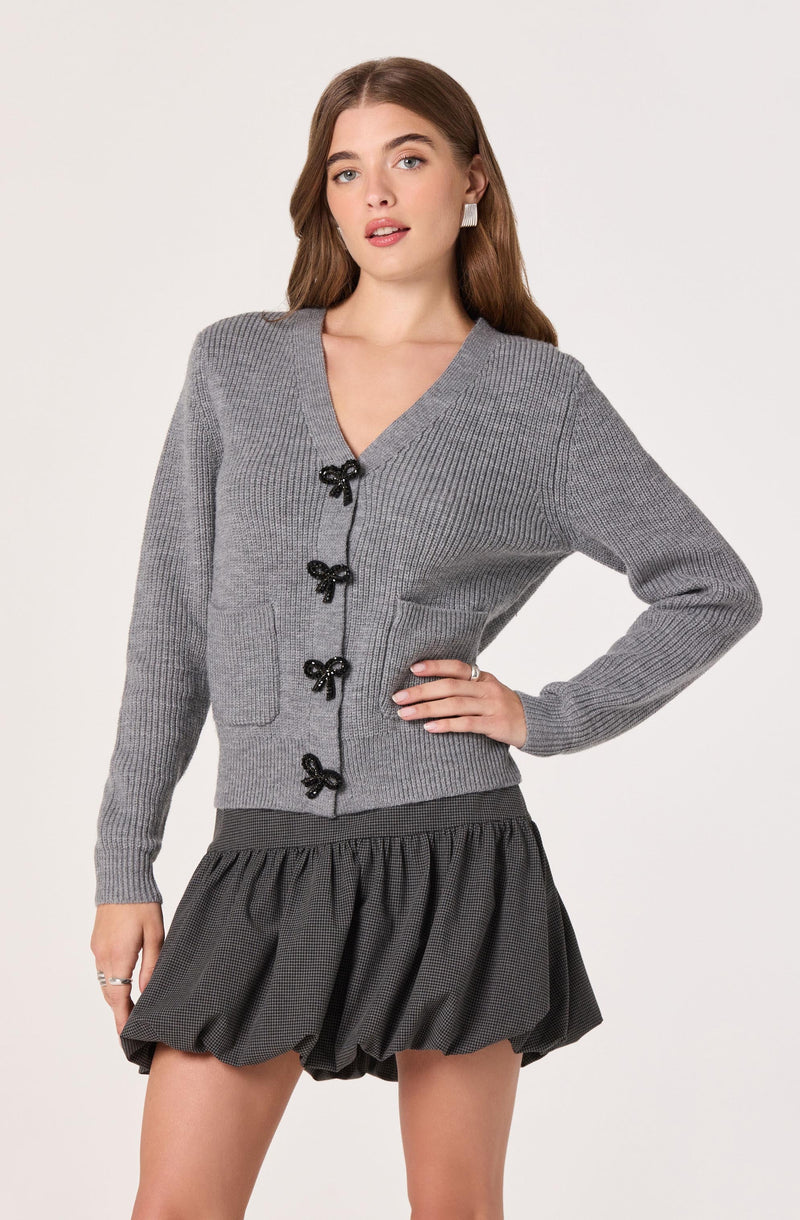 A woman is wearing a light gray, long-sleeved, knit cardigan with a V-neckline and decorative black bow buttons down the front. She has her left hand resting on her hip. She is also wearing a dark, textured, gathered skirt that is above knee length.