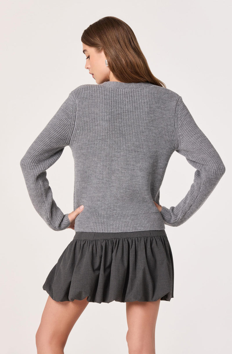 A woman is wearing a grey, long-sleeved, ribbed knit sweater with a crew neckline. She pairs it with a black, ruffled mini skirt that has a gathered, textured fabric.