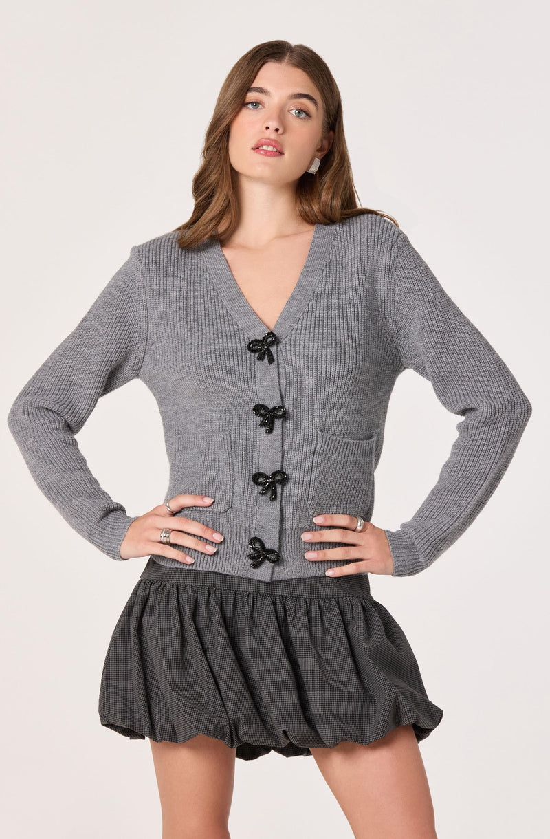 A woman is wearing a light gray, knitted cardigan with a V-neckline and black bow-shaped buttons down the front. The cardigan has long sleeves and a fitted silhouette. She pairs it with a dark, textured, bubble-hem skirt that has an elastic waistband and a gathered, puffy shape.