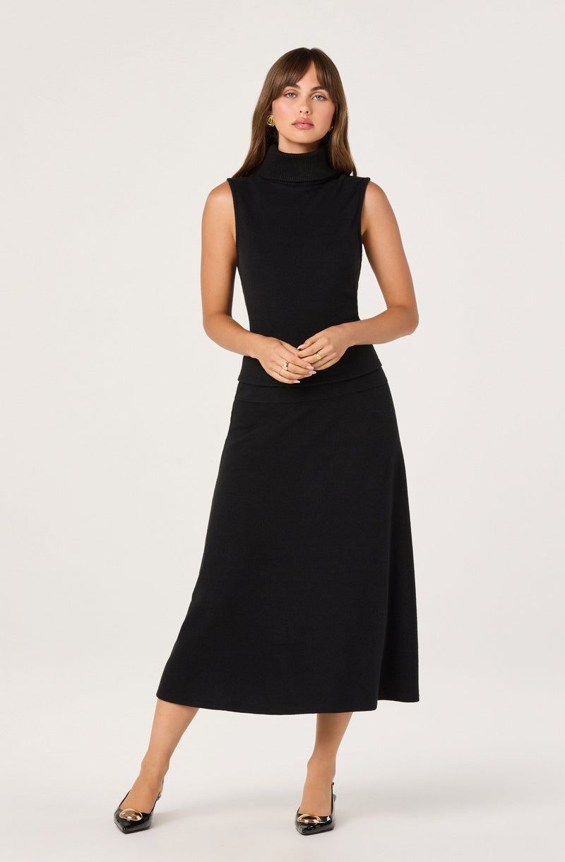 The woman is wearing a sleeveless black turtleneck top and a high-waisted, ankle-length black skirt with a side slit. She is also wearing black pointed-toe heels with gold accents.