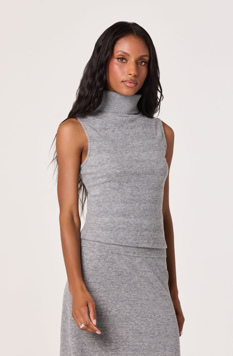 A woman is wearing a sleeveless, high-neck gray knit top with a ribbed collar. The top is fitted and made from a soft, textured fabric. She pairs it with a gray knit skirt that has a matching texture and color, creating a coordinated outfit.