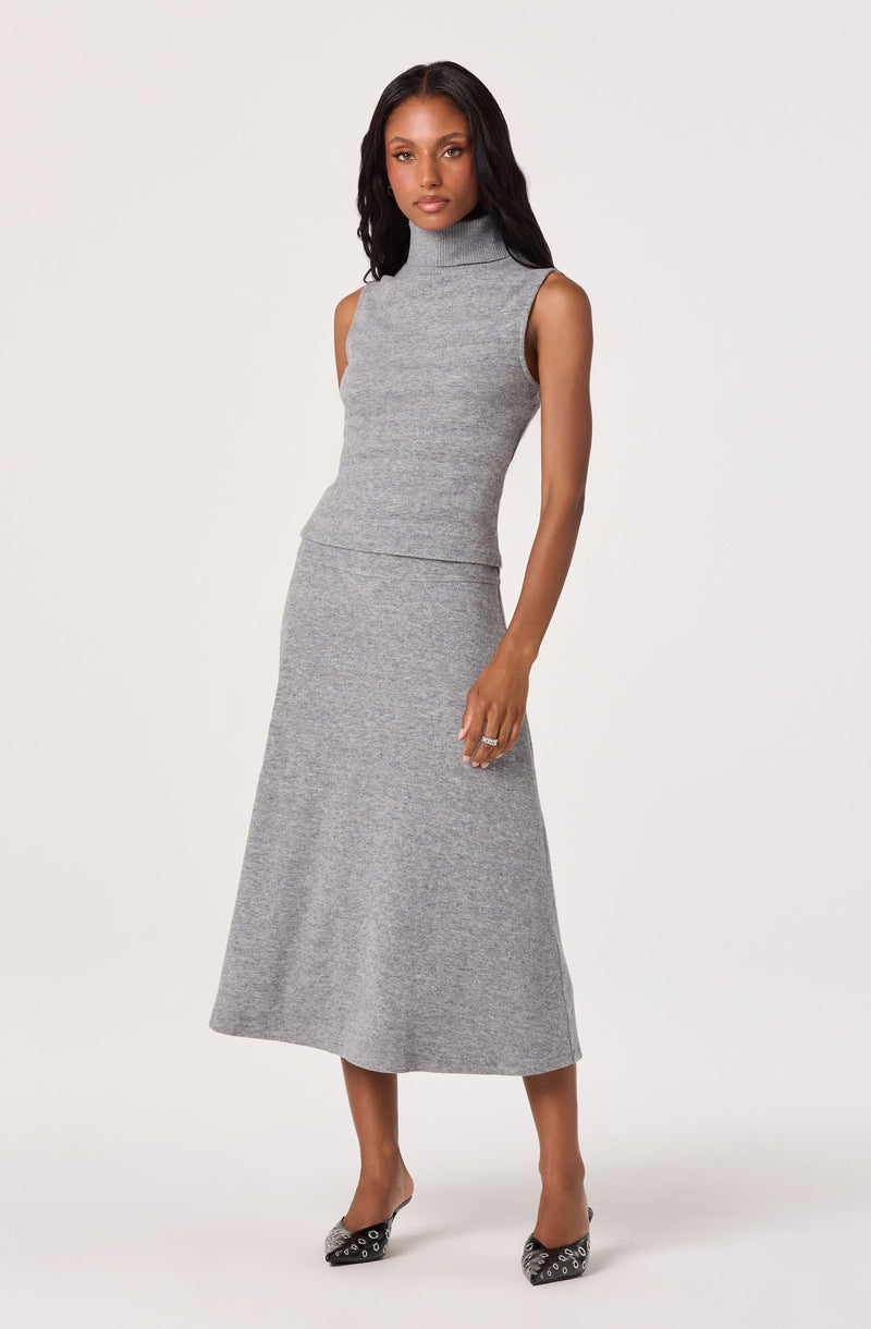 A woman is wearing a sleeveless, high-neck gray knit top paired with a matching gray knit midi skirt. She completes her outfit with black pointed-toe heels decorated with silver eyelet details.