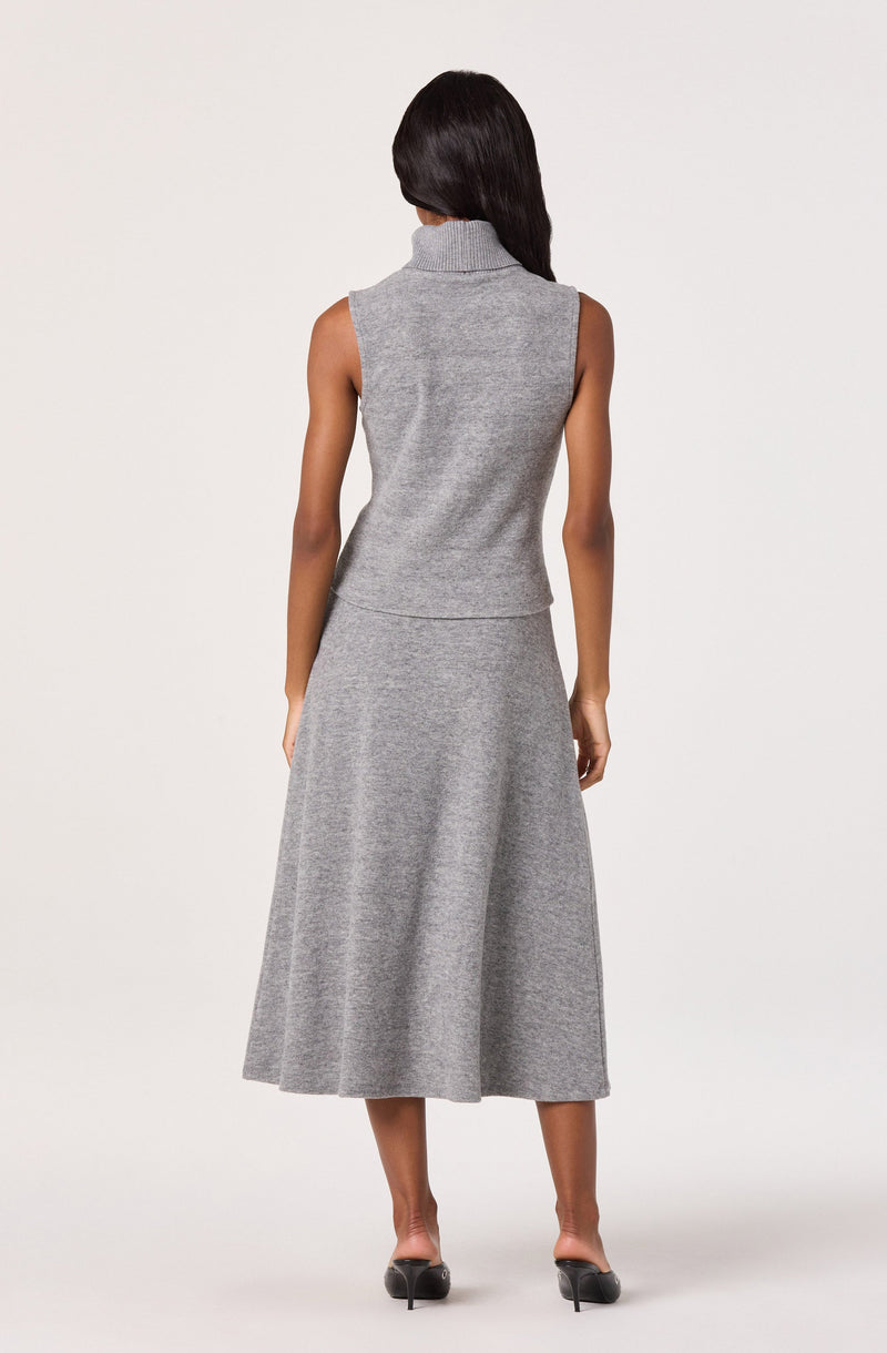 A woman is wearing a sleeveless, gray knit turtleneck top and a matching gray knit A-line midi skirt. She is standing with her back facing the camera on a plain white background.