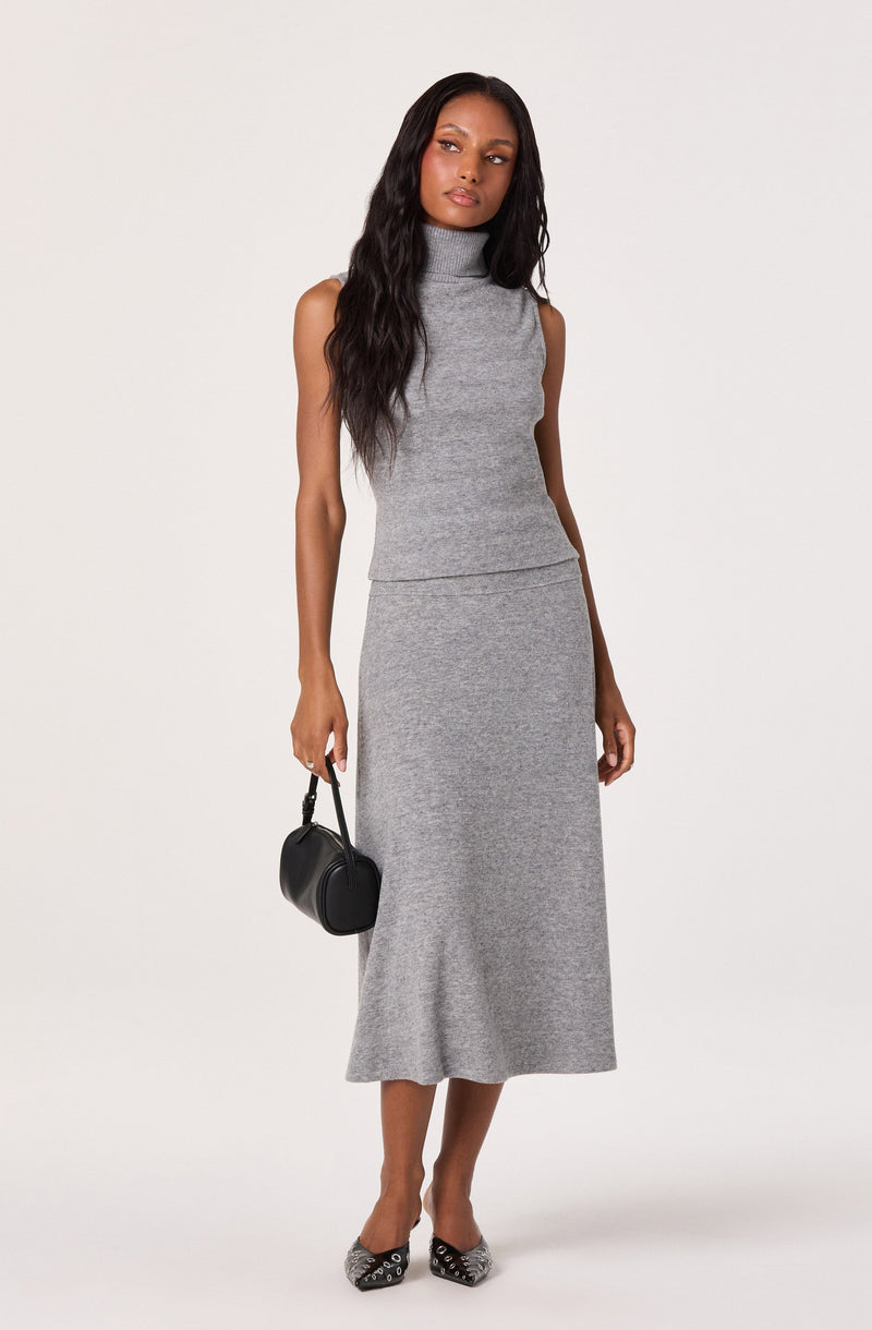 A woman wearing a sleeveless, high-neck, grey knit top paired with a matching long, grey knit skirt. She is holding a small black handbag in her right hand and wearing pointed-toe shoes with a black and white animal print pattern.