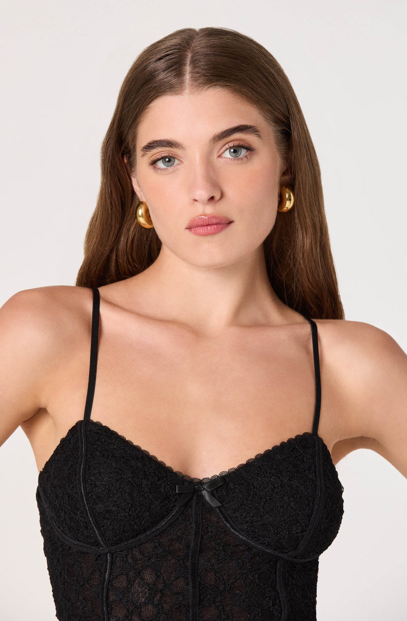 A woman is wearing a black lace camisole with thin spaghetti straps. The camisole features delicate lace fabric with floral patterns and a small bow detail at the center of the bust.