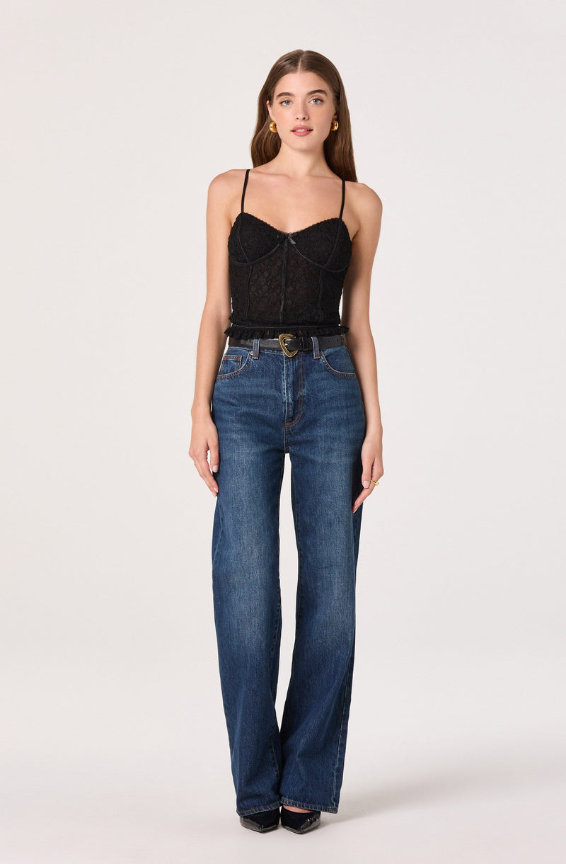 A woman is wearing a black lace camisole top with thin spaghetti straps and a sweetheart neckline. She has paired it with high-waisted, straight-leg blue jeans and black pointed-toe shoes. She accessorizes with gold hoop earrings and a gold ring.