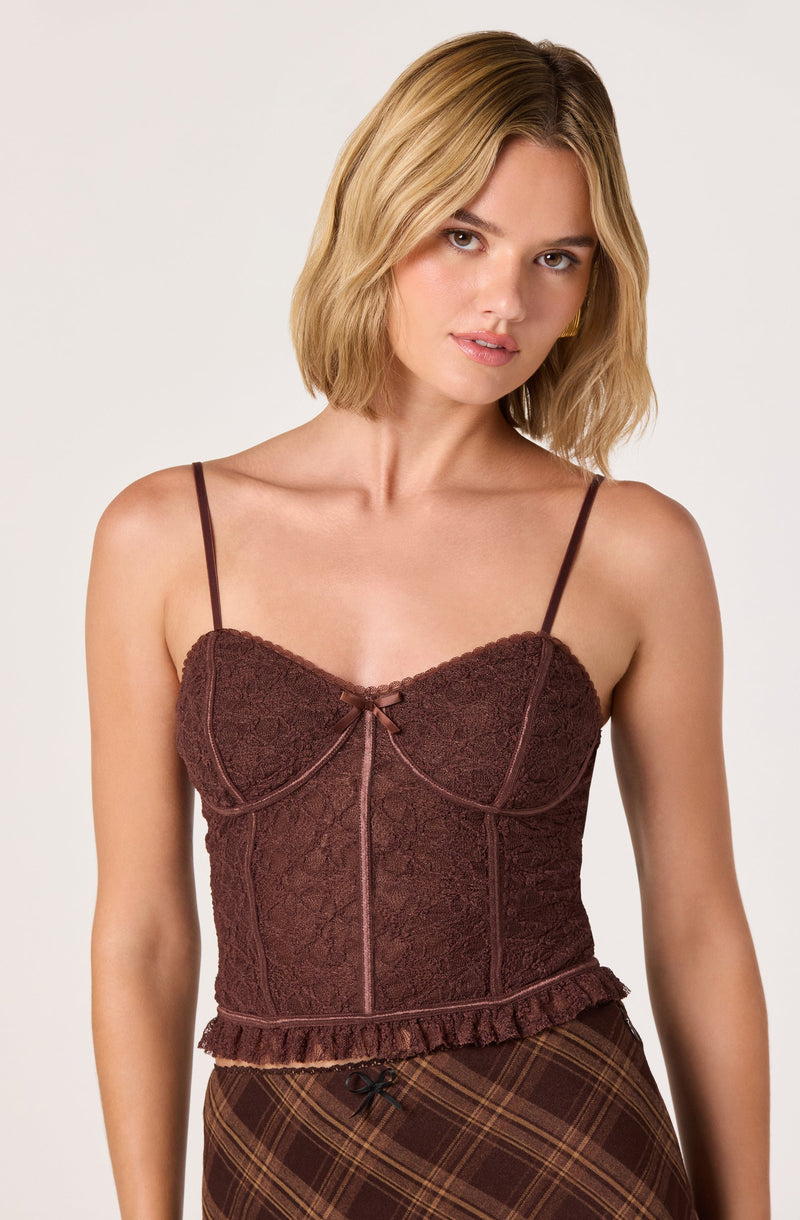 A woman is wearing a fitted, dark brown lace camisole with thin spaghetti straps. The camisole features a small bow at the center of the neckline, vertical stitching details, and a ruffled hem at the bottom. She is also wearing a high-waisted, brown plaid skirt with a small black bow at the waistband.