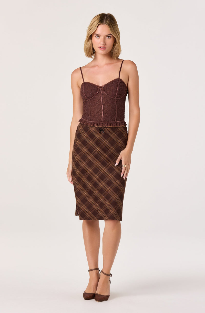 A woman is wearing a fitted, sleeveless top made of textured, reddish-brown fabric with thin spaghetti straps. She pairs it with a high-waisted, knee-length skirt featuring a brown and yellow plaid pattern. She completes her outfit with brown heeled shoes that have ankle straps.