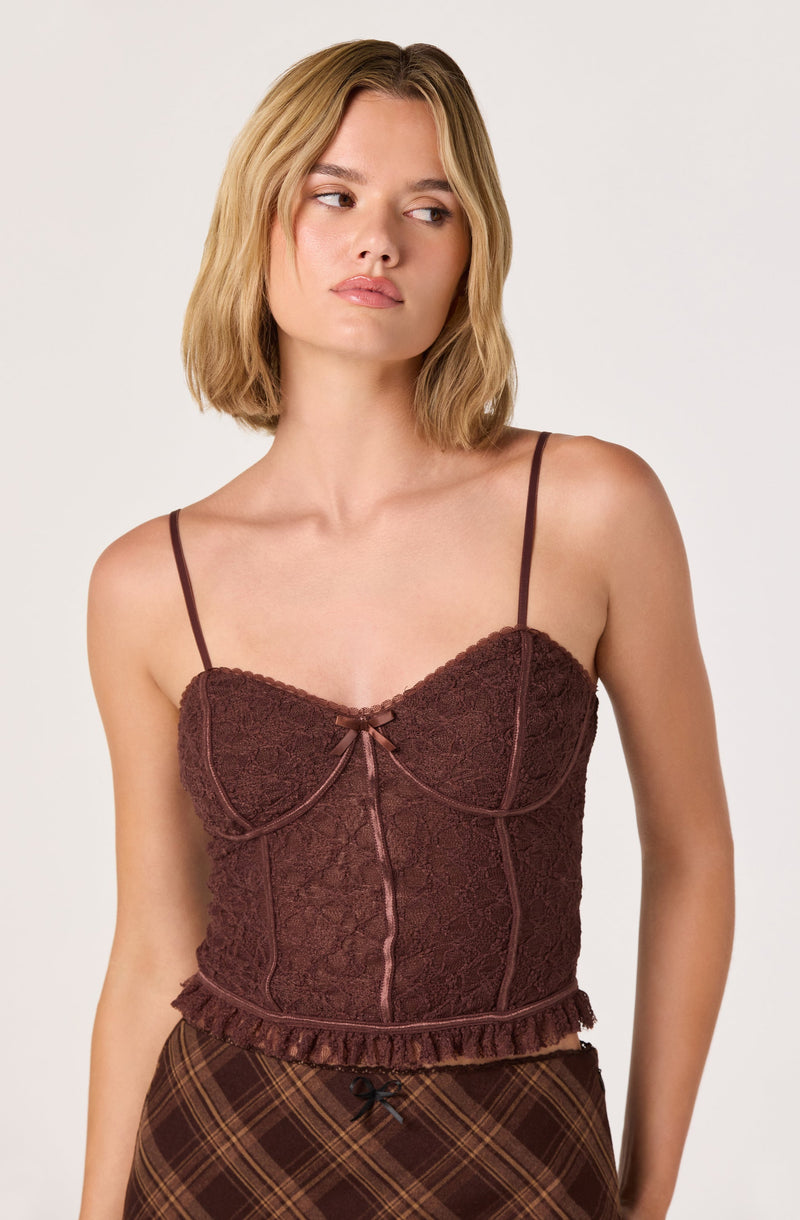 A young woman is wearing a fitted, brown, lace camisole top with thin spaghetti straps. The top features a small bow at the center of the bust and decorative vertical lines running down the front, with a ruffled trim along the bottom hem. The top has a textured lace pattern throughout. She is also wearing a plaid skirt with a dark pattern.