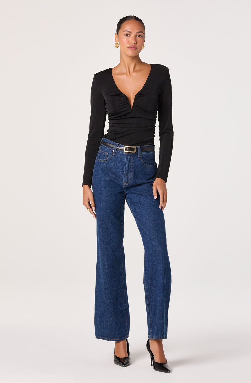 A woman is wearing a fitted black long-sleeve top with a deep V-neckline, ruched detailing at the front, and a smooth texture. She pairs it with high-waisted, wide-leg blue jeans that have a classic five-pocket design and a black belt with a gold buckle. She completes the outfit with black pointed-toe high heels.