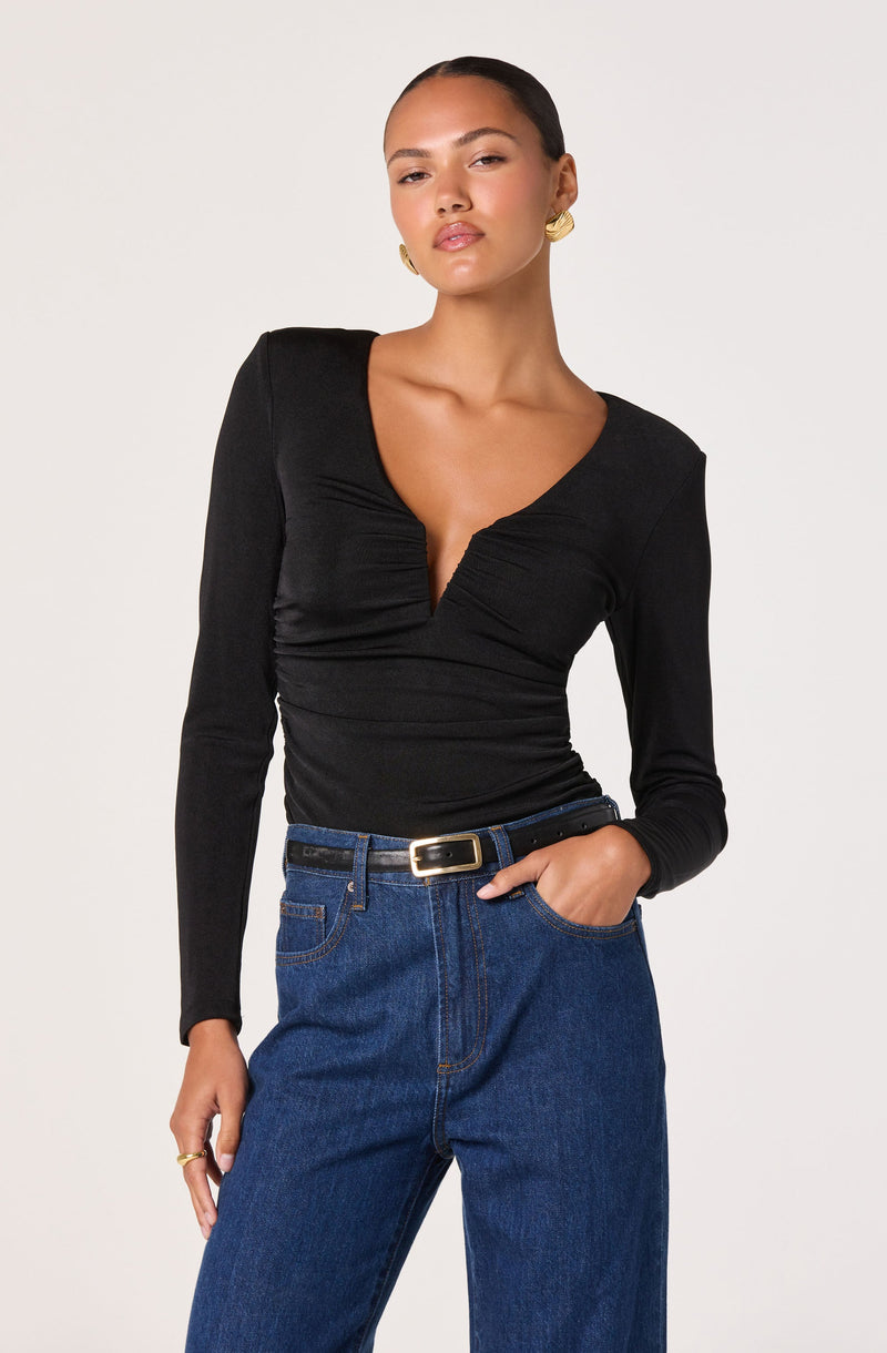 A woman is wearing a fitted black long-sleeve top with a deep V neckline and ruched detailing across the chest, paired with high-waisted blue jeans and a black belt with a gold buckle. She has gold earrings and a gold ring.