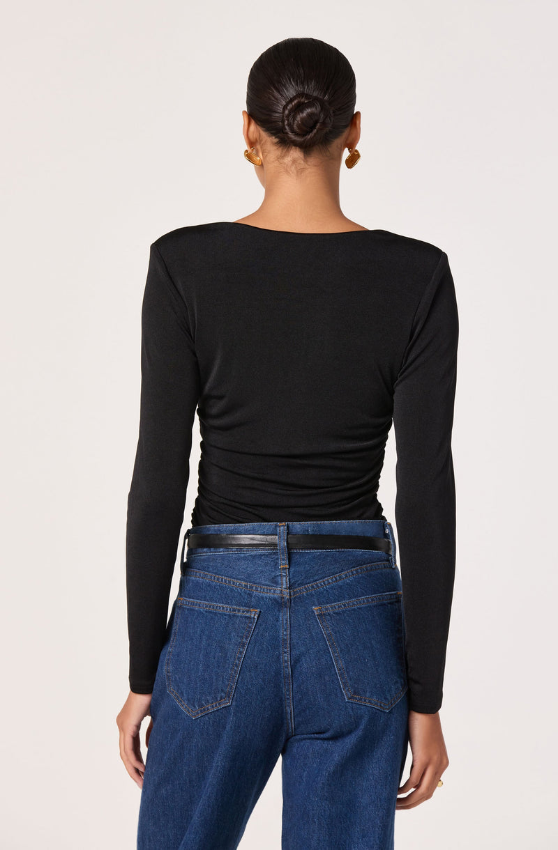 A woman is wearing a fitted black long-sleeve top with a round neckline, paired with high-waisted blue jeans that have visible stitching and back pockets. She is also wearing a black belt.