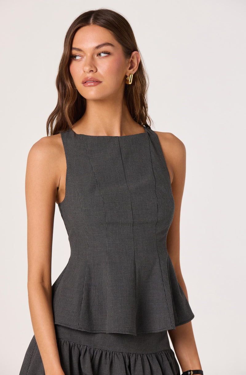 A woman is wearing a sleeveless, fitted, gray and black checkered top with a peplum waist. The top has a broad, rounded neckline and a subtle vertical stripe pattern. She pairs it with a matching skirt that features a ruffled, gathered hem. She has long, wavy brown hair, gold hoop earrings, and a neutral makeup look.