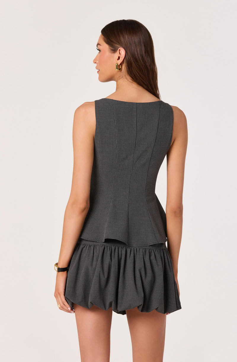 A woman is wearing a sleeveless, fitted grey dress with a textured pattern. The dress has a tailored bodice with a outseam and a peplum detail at the waist. She pairs it with a puffy, gathered skirt that has a bubble silhouette. She accessorizes with gold hoop earrings and a black wristwatch.