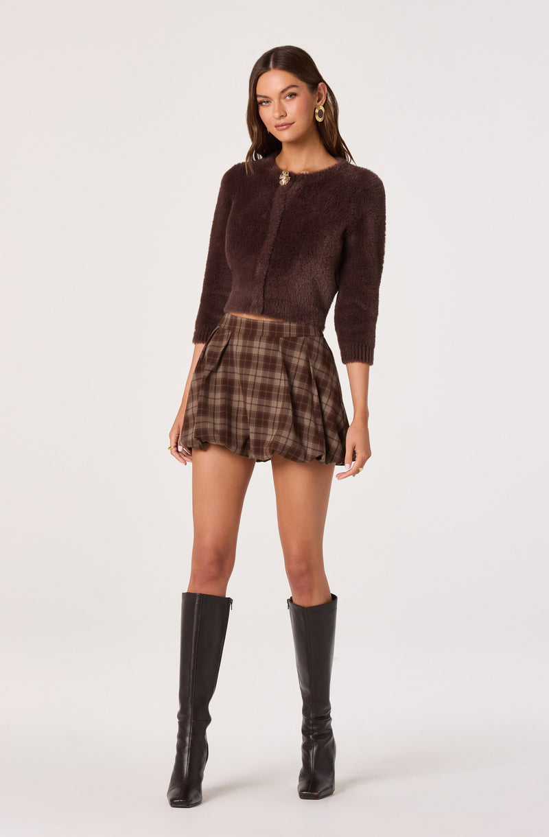 A woman is wearing a dark brown, fuzzy, long-sleeve cardigan with a small, round neckline and a decorative brooch at the top center. She is also wearing a brown and beige plaid pleated mini skirt with a high waist, and black knee-high boots with a slight heel. The overall look is stylish and coordinated with neutral tones.