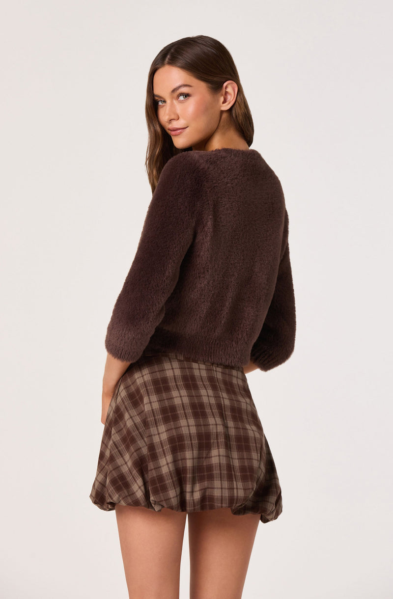 A woman is wearing a dark brown, fuzzy, long-sleeve sweater with a round neckline. She is also wearing a high-waisted, brown plaid skirt with a gathered, slightly puffed hem that creates a ruffled appearance.