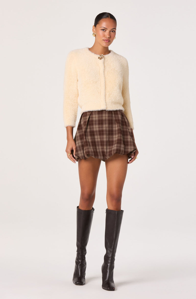A woman is wearing a textured, cream-colored, cropped cardigan with a decorative brooch at the neckline, paired with a brown plaid skirt that has a slightly puffed, bubble-style silhouette. She is also wearing tall black leather boots that reach just below her knees.