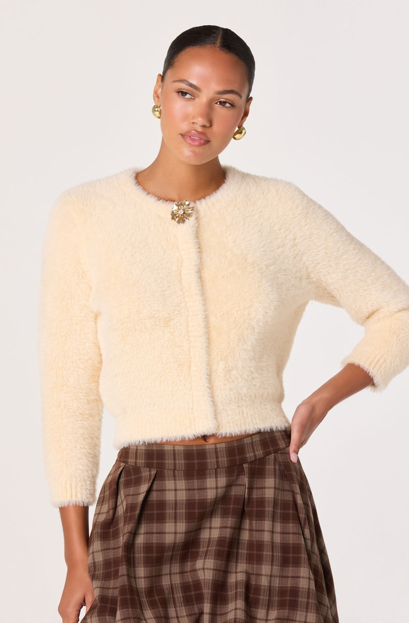 The woman is wearing a soft, cream-colored, cropped cardigan with a fuzzy texture. The cardigan has a round neckline decorated with a flower-shaped brooch. She is also wearing a brown plaid skirt with pleats. Her accessories include gold, circular earrings.