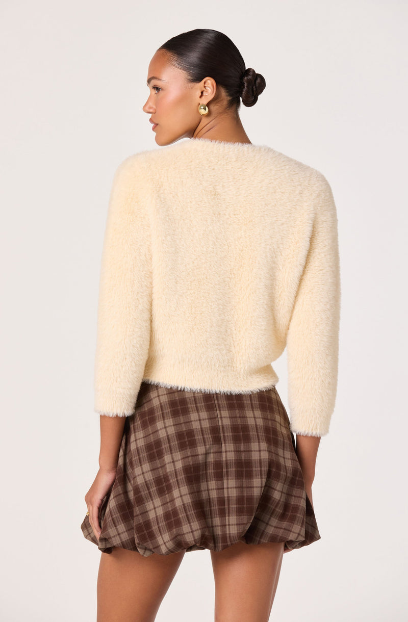 A woman is wearing a soft, fuzzy cream-colored sweater with three-quarter length sleeves and a relaxed fit. She pairs it with a high-waisted, brown plaid skirt featuring a puffy, bubble hem that creates a voluminous silhouette. Her hair is styled in a sleek, low bun, and she is accessorized with large, gold-toned earrings.