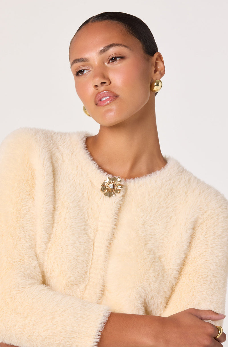A woman wearing a light cream, fluffy, textured sweater with a round neckline. The sweater has a decorative large metallic flower-shaped clasp at the top. She is also wearing bold gold earrings.
