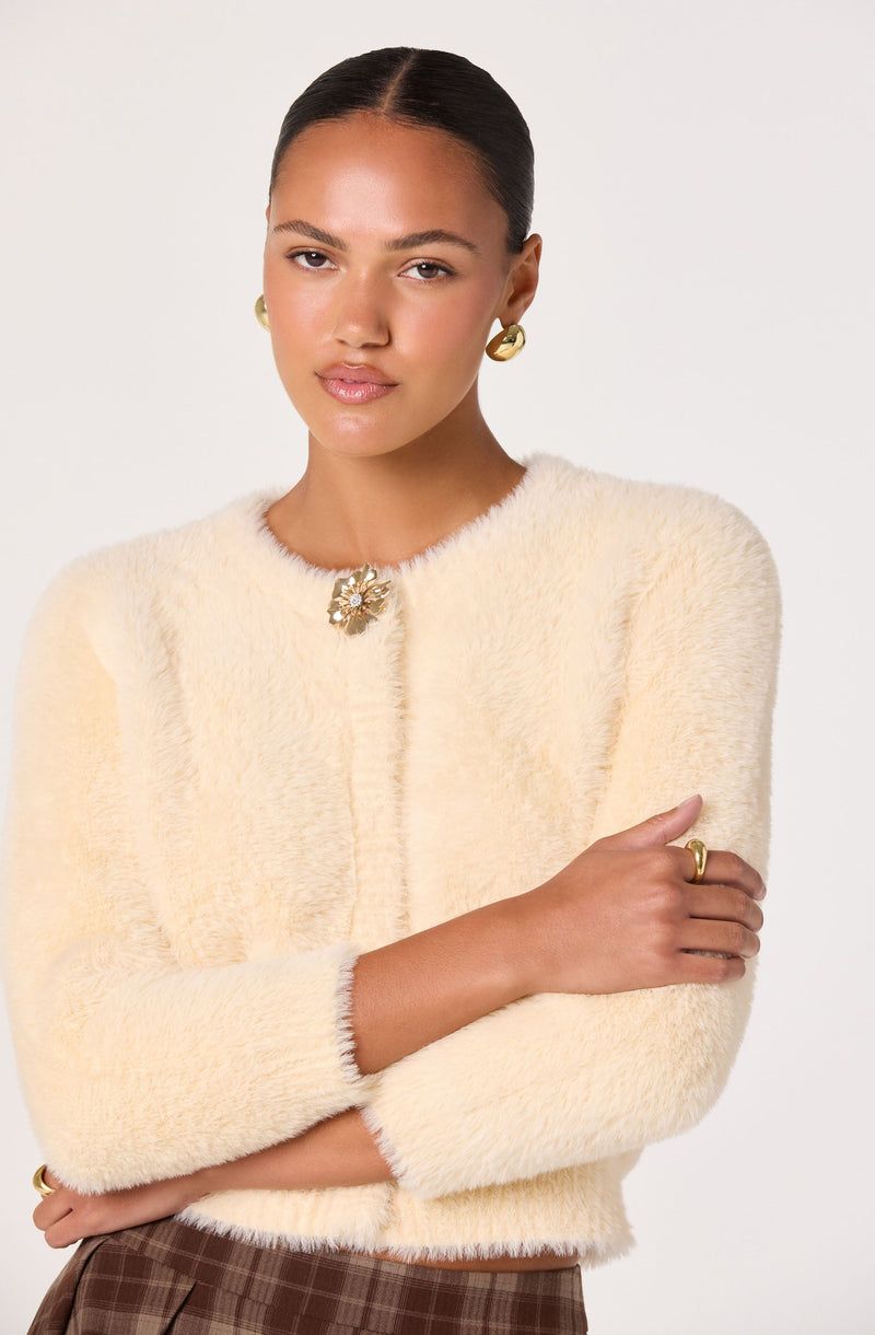 A woman wearing a soft, cream-colored, fuzzy textured long-sleeve jacket with a decorative floral pin at the neckline. She has on gold hoop earrings and a gold ring, and her arms are crossed.
