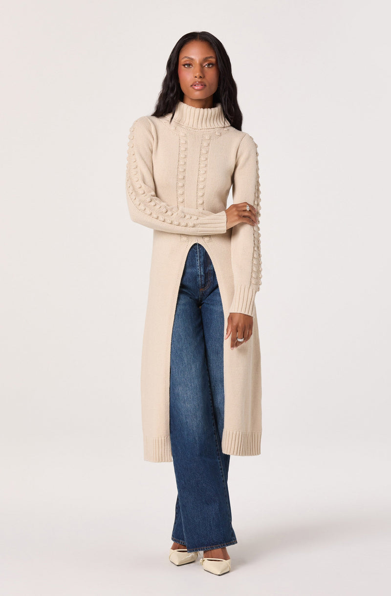 A woman is wearing a long, chunky knit cream-colored sweater with a ribbed turtleneck and intricate cable knit patterns run down the front and along the sleeves. The sweater has side slits that reveal high-waisted, wide-leg blue jeans. She completes the look with pointed cream-colored heels.