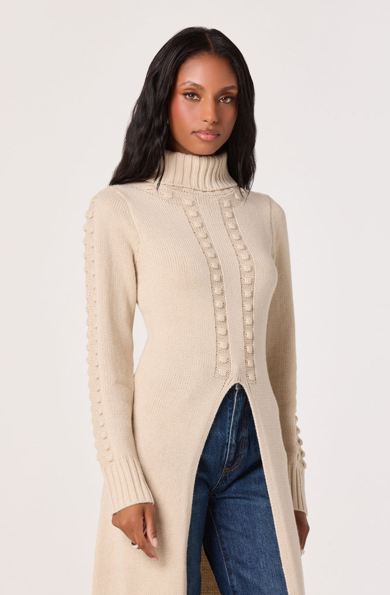A woman is wearing a long, cream-colored knit cardigan with a high ribbed turtleneck. The cardigan features a textured pattern with raised rectangular details running vertically along the front and along the length of the sleeves. The cardigan is open in the front, revealing a pair of dark blue jeans.