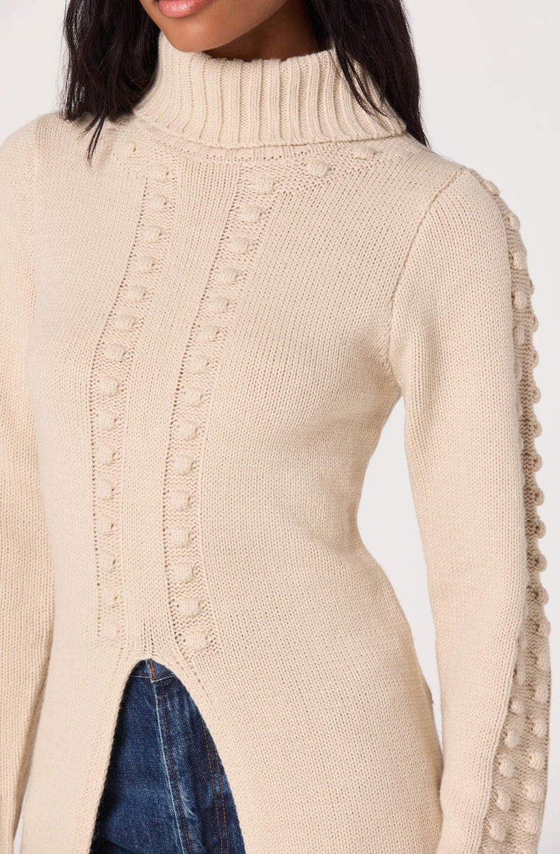 A woman is wearing a beige knit sweater with a high ribbed turtleneck collar. The sweater features textured vertical cable knit patterns and decorative bobble details along the sleeves and front. The material appears thick and cozy. She is also wearing blue jeans.