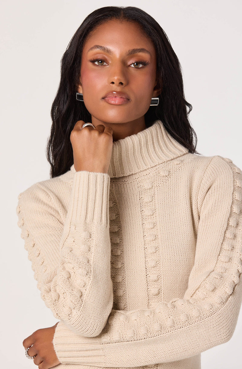 A woman is wearing a cream-colored, chunky knit turtleneck sweater with detailed textured patterns along the sleeves and front. The sweater has a high ribbed collar and features raised knit designs running vertically down the center and along the sleeves.