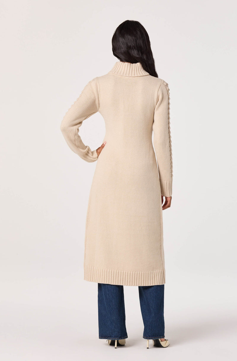 A woman is wearing a long, cream-colored knit cardigan with a high collar. The cardigan features textured detail along the sleeves and has a ribbed hem. She pairs it with dark blue jeans and black-and-white high-heeled shoes.