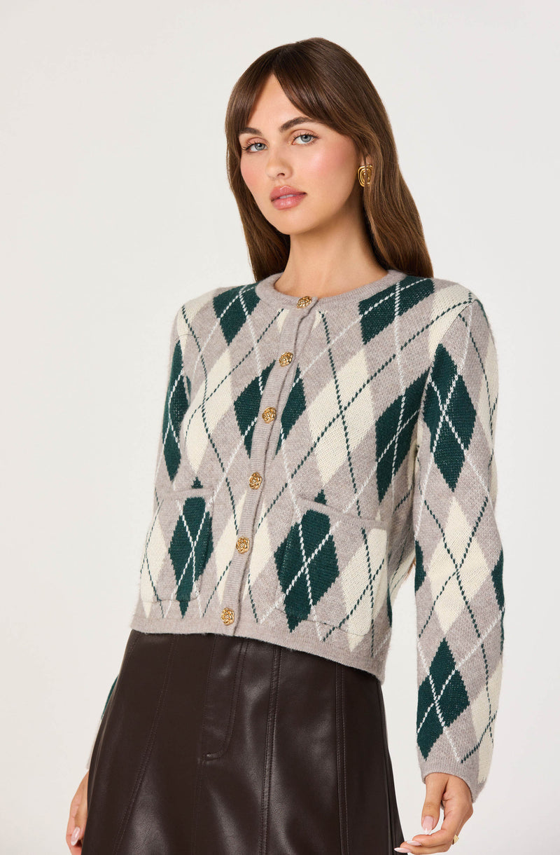 A woman is wearing a cropped cardigan with an argyle pattern featuring large dark green diamonds with white and light gray lines on a beige background. The cardigan has a round neckline and gold buttons down the front.