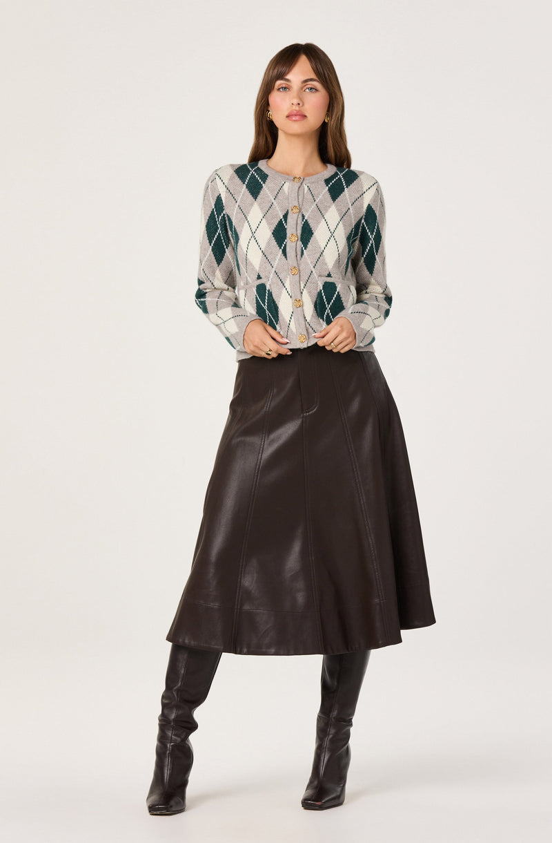 The woman is wearing a light-colored button-up cardigan with an argyle pattern in shades of green, beige, and white. She pairs it with a dark brown, leather A-line skirt that falls below the knees. She completes the look with tall, dark leather boots that reach just above the knees.