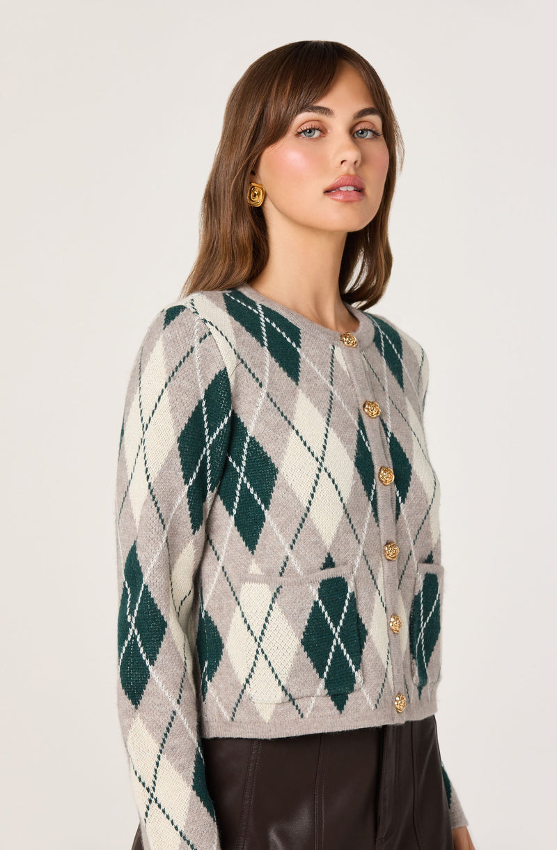 A woman is wearing a light gray cardigan with an argyle pattern of dark green, beige, and white diamonds. The cardigan has a round neckline and is buttoned with gold decorative buttons. She is also wearing gold earrings.
