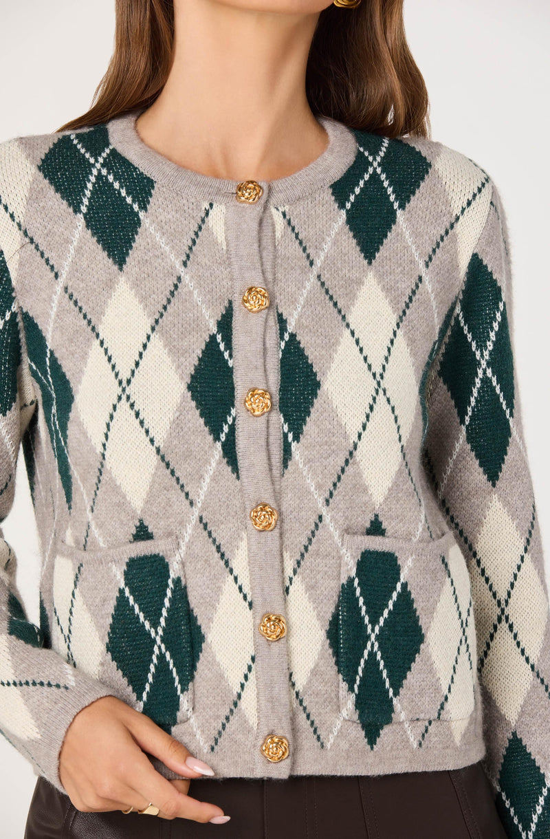 A woman is wearing a cardigan with a classic argyle pattern. The cardigan features a light gray base color with large, dark green diamonds and smaller, cream-colored diamonds intersected by thin, dark green lines. It has a round neckline and is fastened down the front with gold, floral-shaped buttons. The cardigan also has two front pockets near the waist, aligned with the diamond pattern.