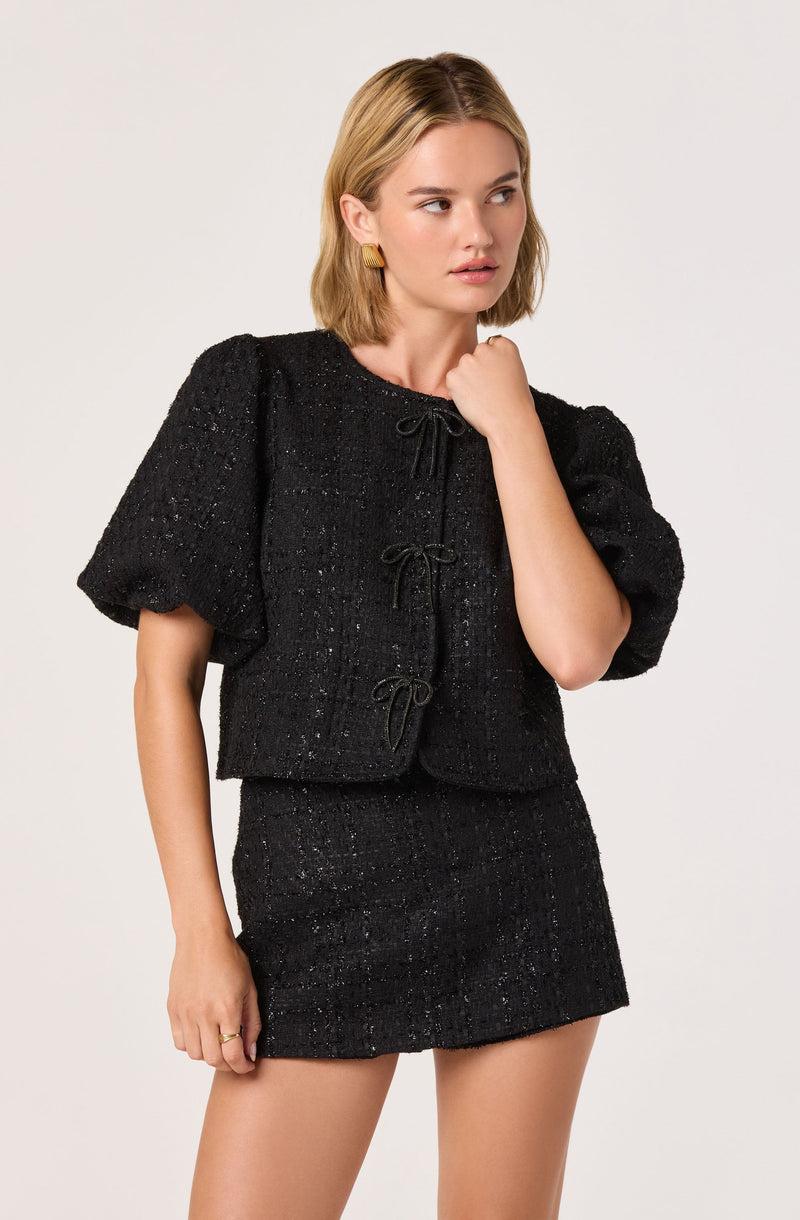 A woman is wearing a black textured short-sleeve jacket with puffed sleeves and a matching black textured mini skirt. The jacket features a round neckline and is fastened with three small bows down the front.