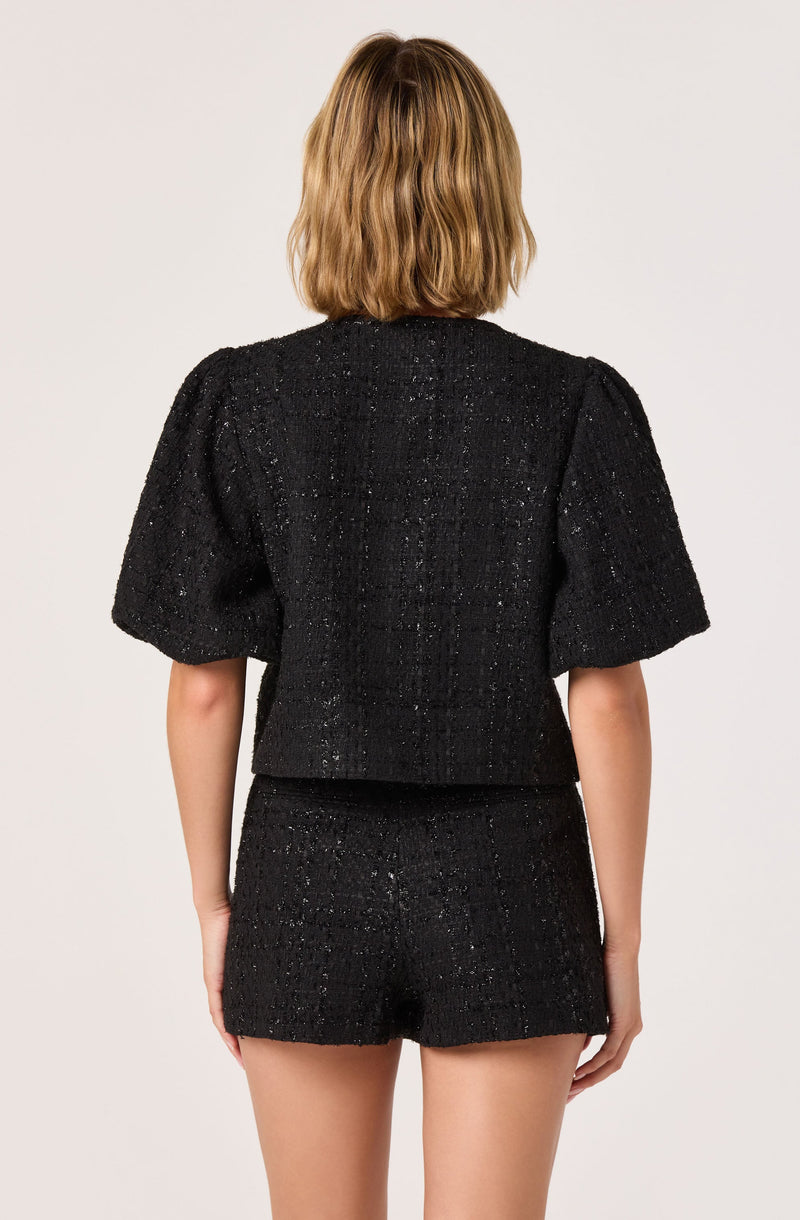 A woman is wearing a black textured two-piece outfit consisting of a short-sleeved top with puff sleeves and matching high-waisted shorts. The fabric appears to have a slightly shiny, speckled finish.