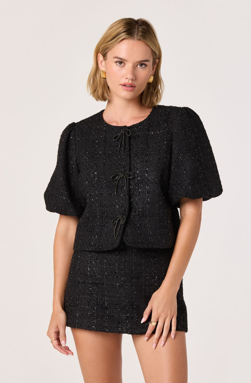 A woman is wearing a black textured two-piece outfit consisting of a short-sleeved, cropped jacket with puff sleeves, and a matching high-waisted mini skirt. The jacket features a round neckline and is fastened with three decorative bows down the front. The fabric has a slightly shimmery, woven appearance.