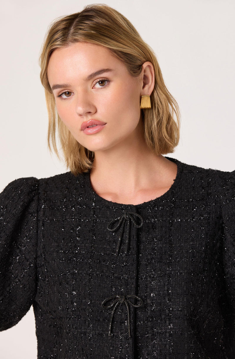 A woman with shoulder-length blonde hair wearing a textured black top with a round neckline. The top features a subtle sparkle and has decorative bows at the front with thin black ties. She is accessorized with gold, rectangular earrings.