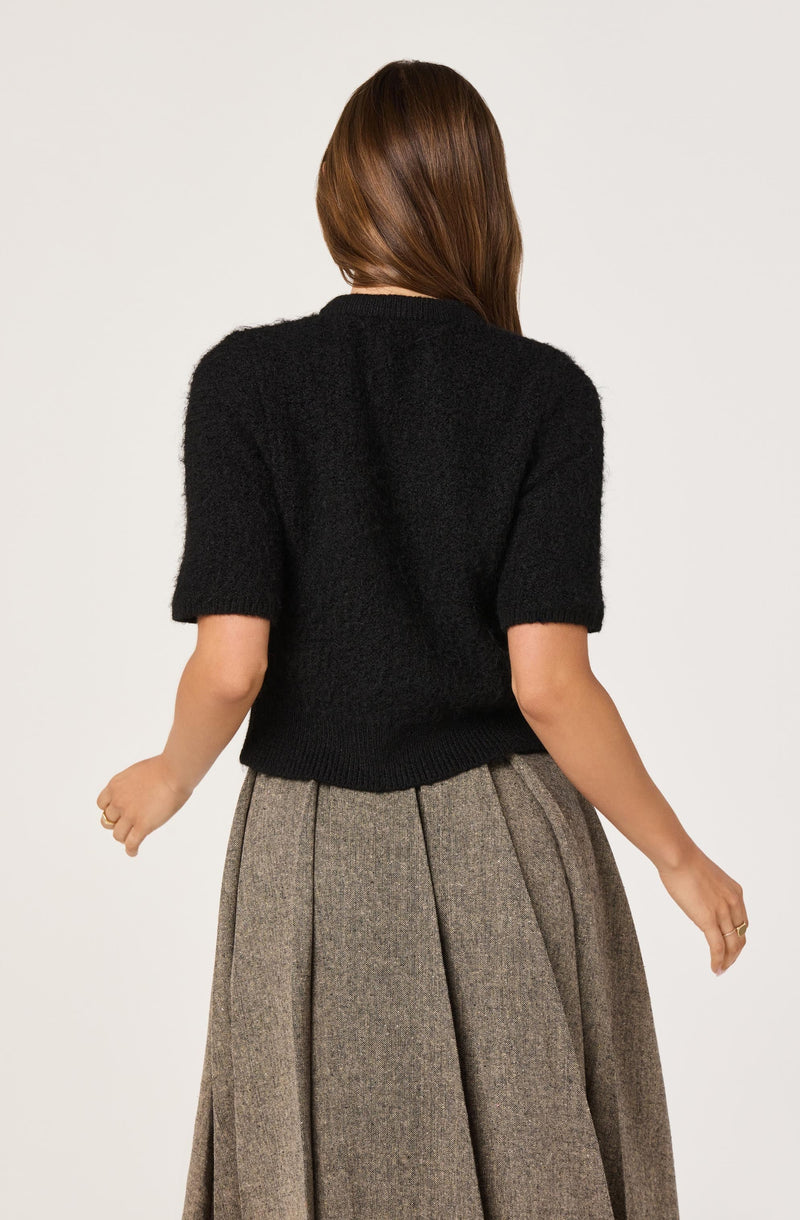 A woman is wearing a black, short-sleeved, fuzzy knit sweater and a high-waisted, pleated, gray plaid skirt.