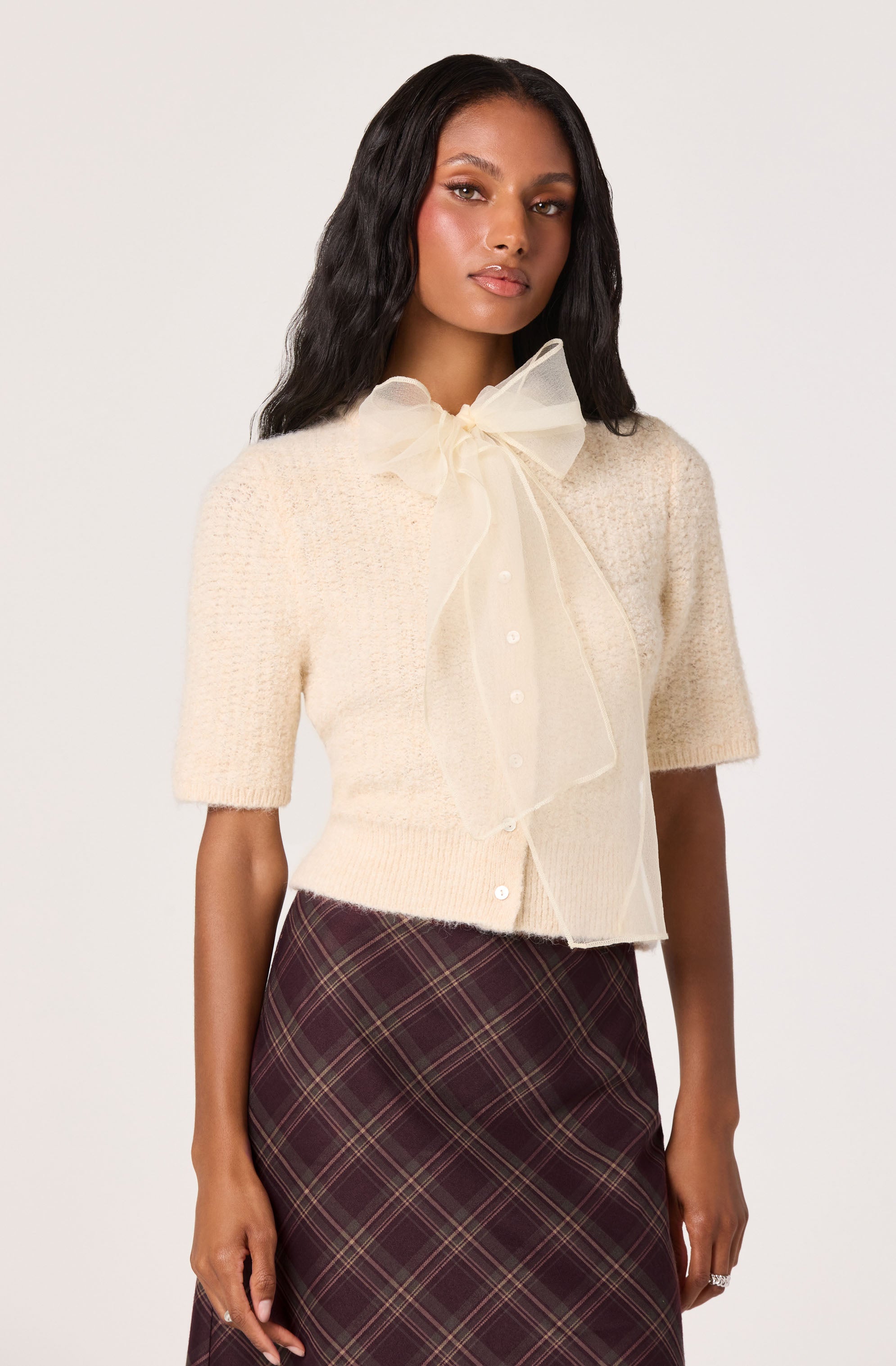 A woman is wearing a short-sleeved, cream-colored textured cardigan with a large, sheer bow tie at the collar. She pairs it with a high-waisted, dark plaid skirt featuring a pattern of intersecting lines in shades of red, gold, and cream.