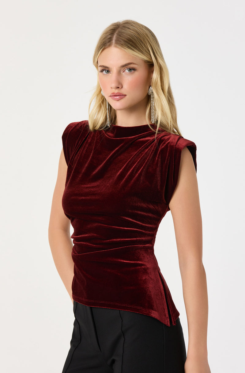 A woman is wearing a sleeveless, deep red velvet top with a high neckline and subtle draping details across the front. She has long, wavy blonde hair and is accessorized with long, dangling earrings.