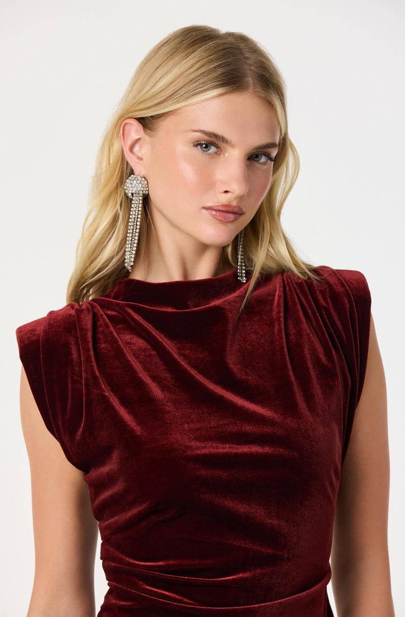 A woman is wearing a sleeveless, deep red velvet dress with a high, rounded neckline. The dress features gathered fabric at the shoulders, creating a slightly puffed sleeve effect. She is also wearing long, dangling earrings that are encrusted with gemstones.