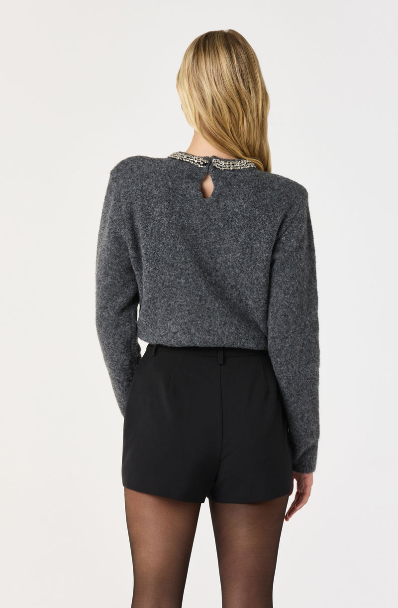 The image shows a woman wearing a dark gray, long-sleeved sweater with a beaded embellishment around the neckline and a small keyhole opening at the back. She is also dressed in high-waisted black shorts and sheer black tights.