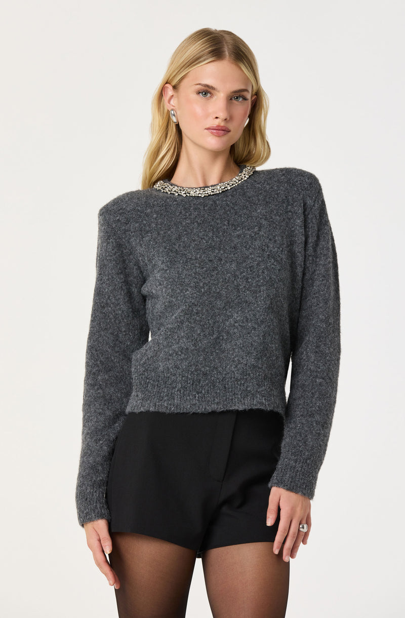 A woman is wearing a dark grey, long-sleeve sweater with a decorative, beaded collar. She pairs it with black shorts and sheer black tights.