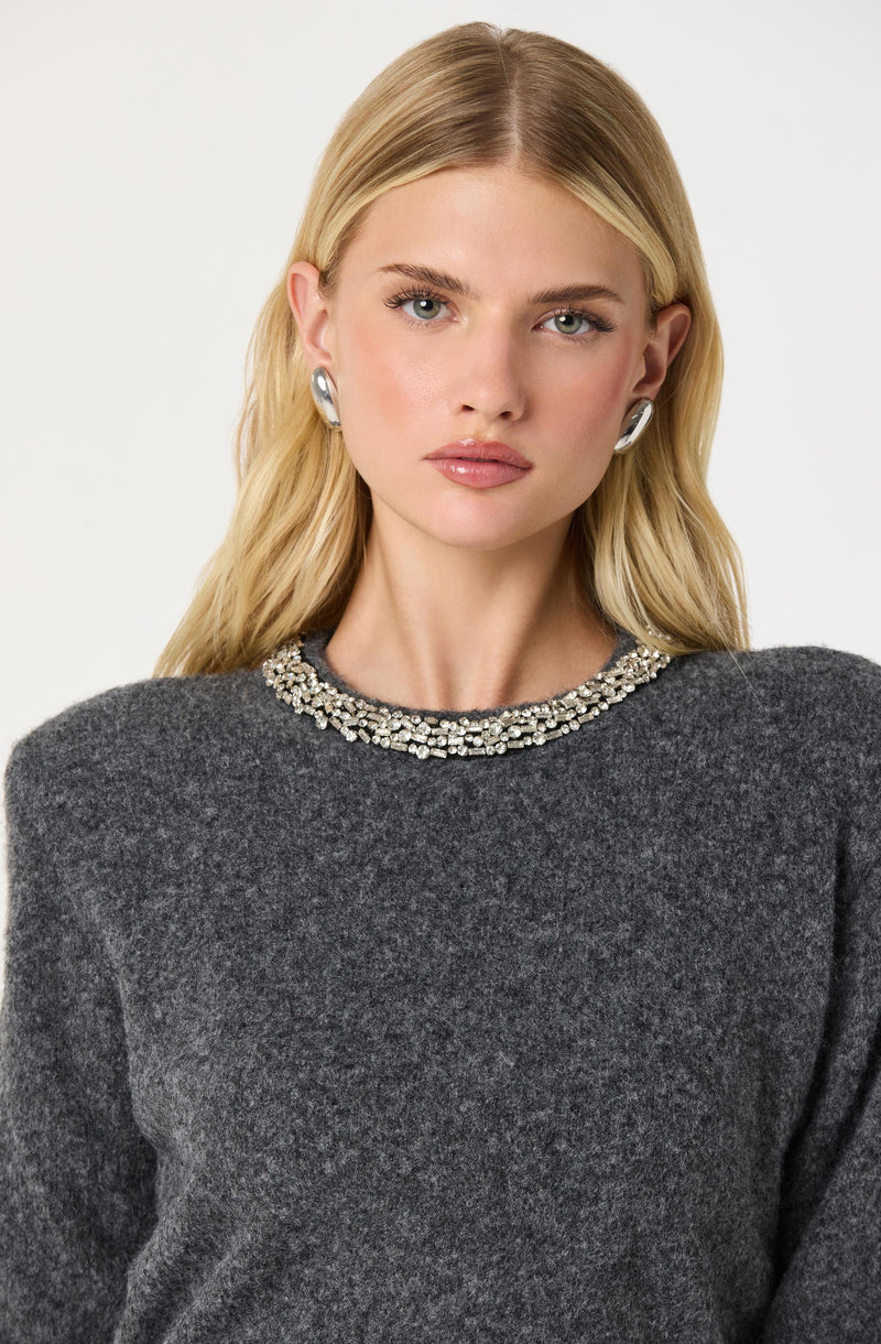 A woman with blonde hair wearing a dark gray sweater with a high neckline, embellished with a beaded or jeweled collar. She is also wearing large, shiny, silver hoop earrings.